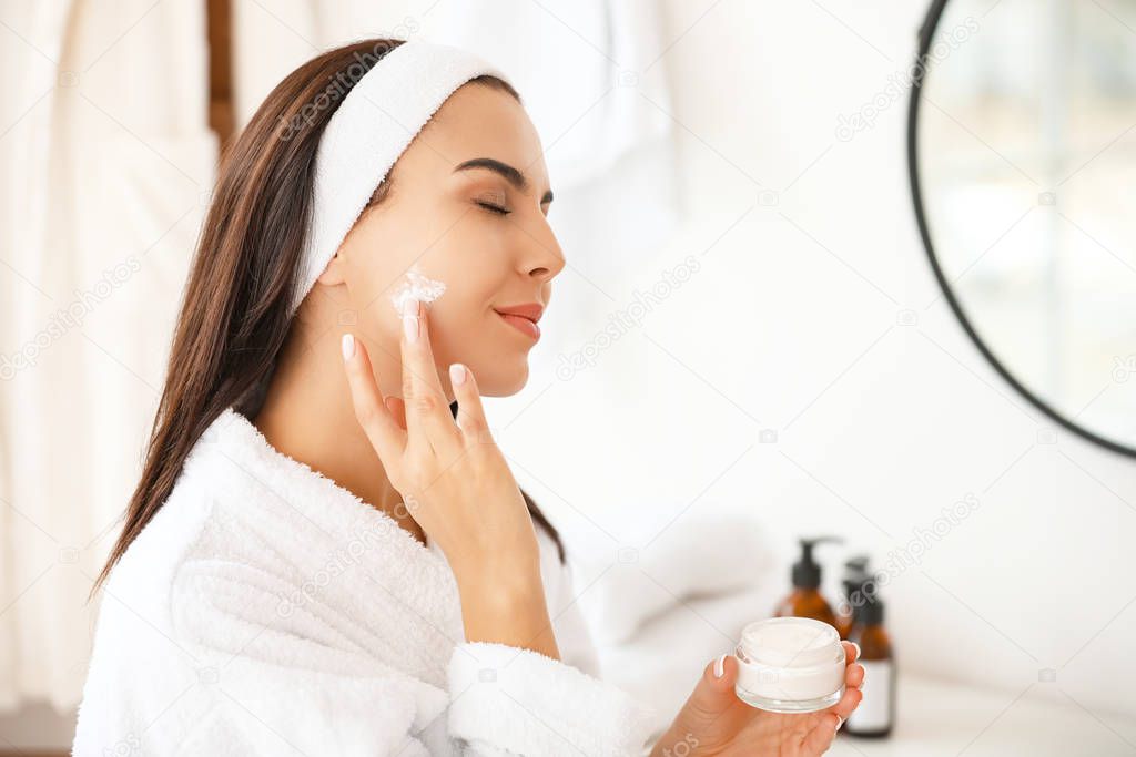 Beautiful young woman applying cream in bathroom