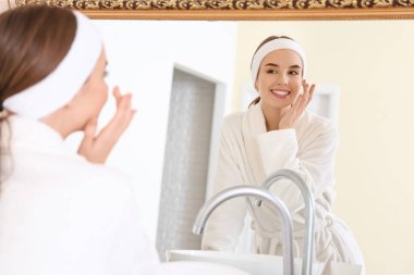 Güzel genç bir kadın banyoda aynanın yanında yüzüne krem sürüyor.