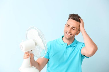 Young man with electric fan on color background