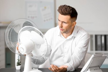 Confused man suffering from heat near electric fan in office