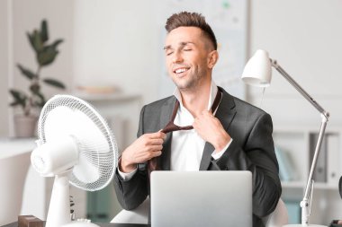 Young man using electric fan during heatwave in office