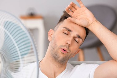 Young man using electric fan during heatwave at home