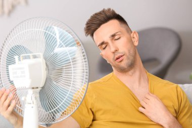 Young man using electric fan during heatwave at home