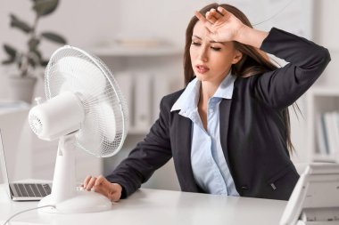 Ofiste sıcak hava dalgası sırasında elektrikli yelpaze kullanan genç bir kadın.