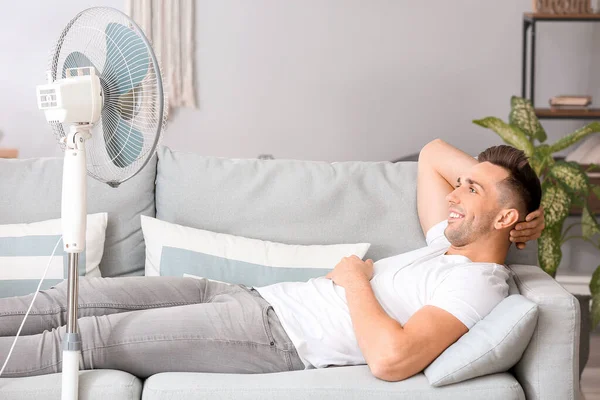 Hombre joven usando ventilador eléctrico durante la ola de calor en casa — Foto de Stock