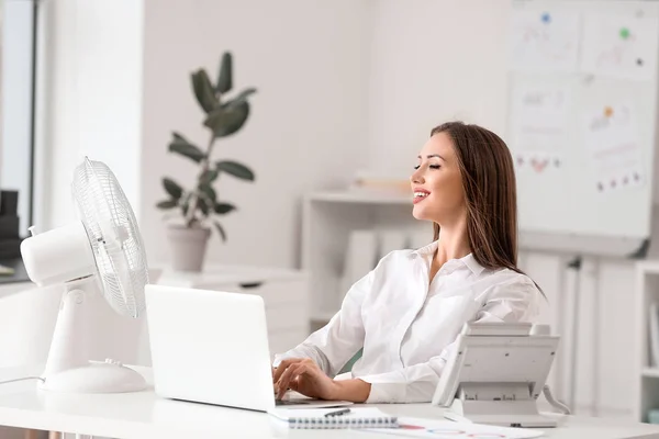 Young woman using electric fan during heatwave in office