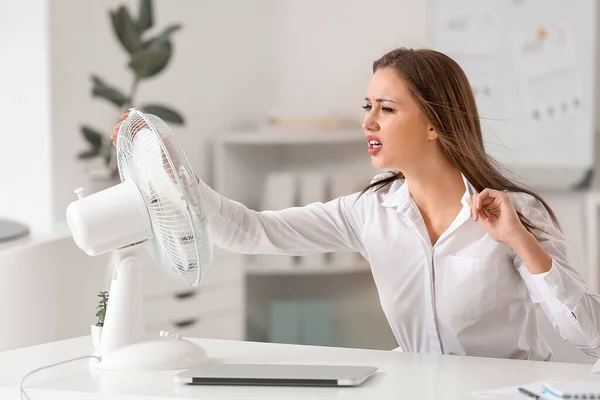 Indignant woman suffering from heat near electric fan in office — Stock Photo, Image