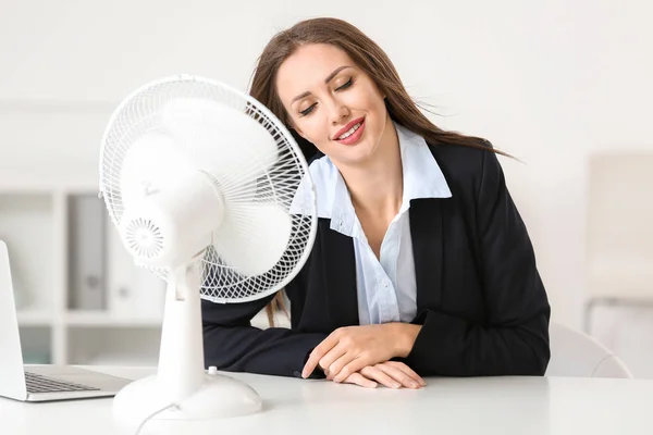 Young woman using electric fan during heatwave in office — Stock Photo, Image