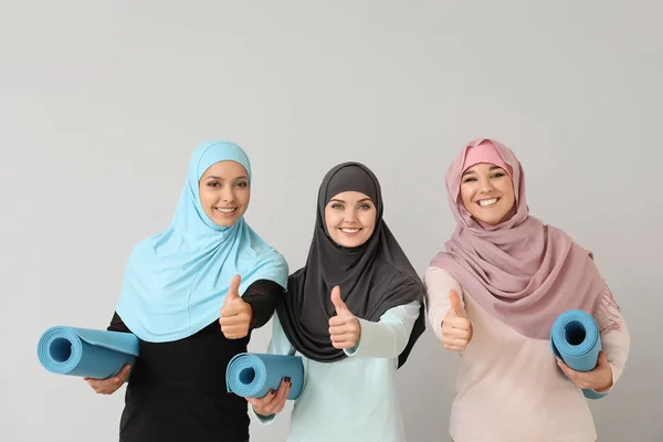 Sportieve moslim vrouwen met yoga matten tonen duim-up op lichte achtergrond — Stockfoto