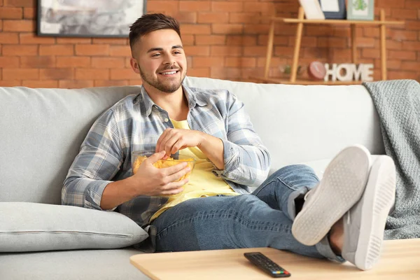 Handsome young man eating tasty potato chips while watching TV at home — Stock Photo, Image