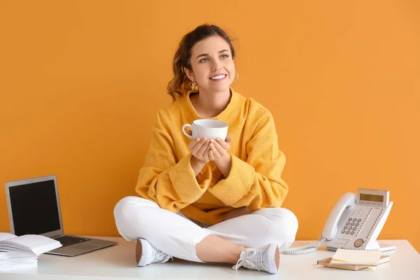 Beautiful young woman with cup of tea sitting on table against color background — 스톡 사진