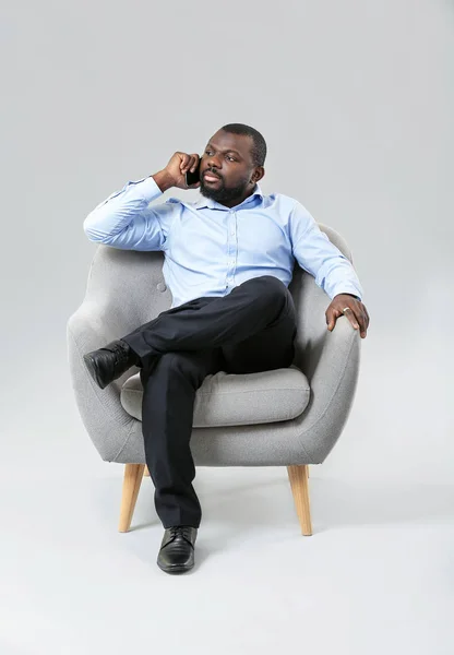 African-American man talking by phone while sitting in armchair against grey background — Stock Photo, Image