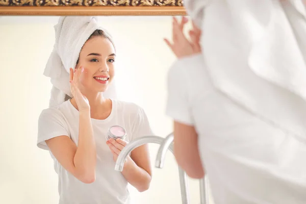 Mulher bonita aplicando creme no rosto perto do espelho no banheiro — Fotografia de Stock