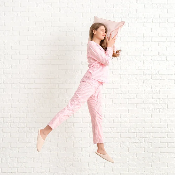 Jumping young woman in pajamas and with pillow on white brick background — 스톡 사진