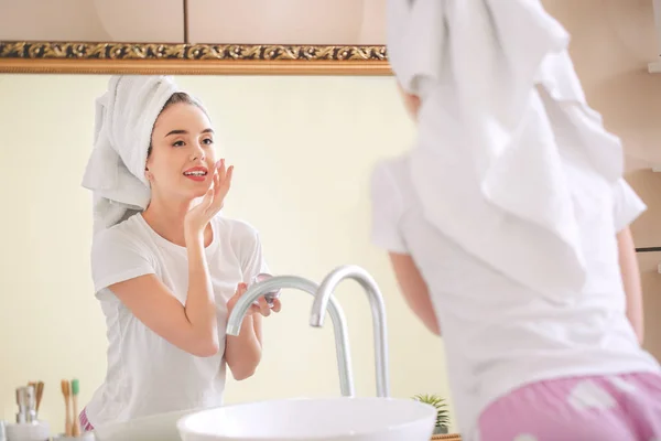 Beautiful young woman applying cream on her face near mirror in bathroom — Stock Photo, Image