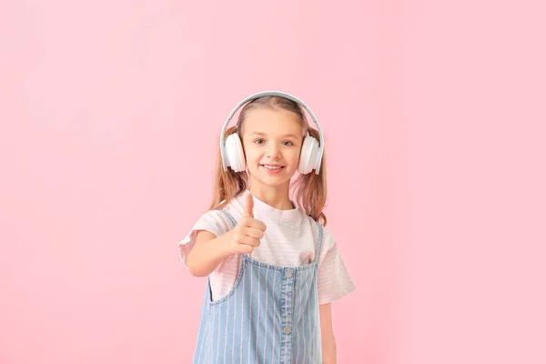 Cute little girl with headphones showing thumb-up gesture on color background — Stock Photo, Image