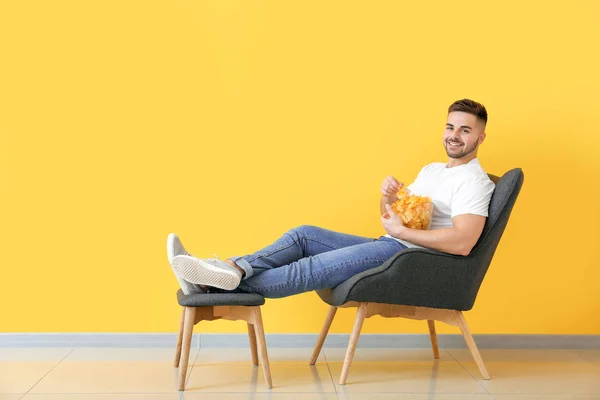 Handsome young man with tasty potato chips sitting in armchair near color wall — Stock Photo, Image