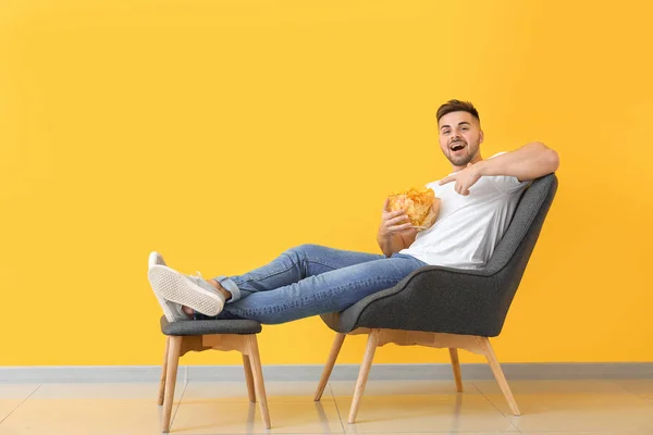 Handsome young man with tasty potato chips sitting in armchair near color wall — Stock Photo, Image