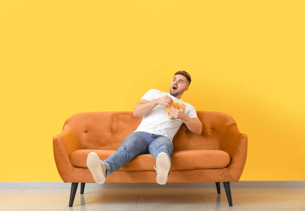 Scared young man with tasty potato chips sitting on sofa near color wall — Stock Photo, Image