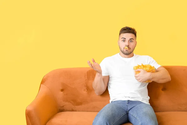 Joven sorprendido con sabrosas papas fritas sentado en el sofá cerca de la pared de color — Foto de Stock