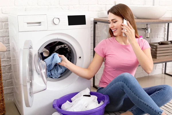 Young housewife talking by phone while washing laundry at home — 스톡 사진