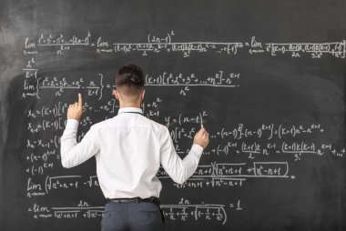 Sınıfta tahtaya yakın bir yerde işaret parmağı kaldırılmış bir erkek öğretmen.