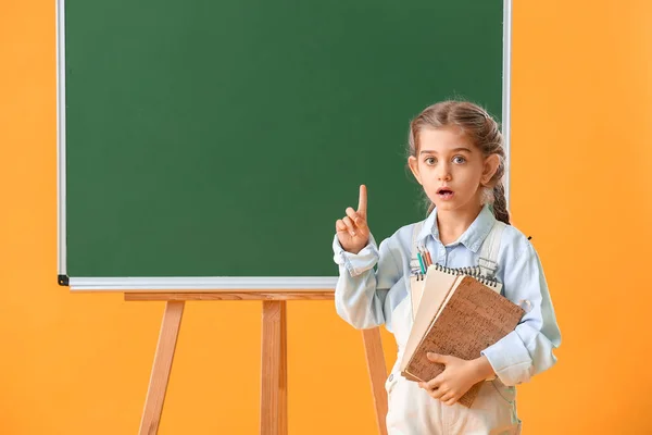 Mała uczennica z podniesionym palcem wskazującym w pobliżu tablicy na tle koloru — Zdjęcie stockowe