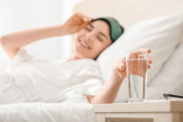 Morning of beautiful young woman taking glass of water from bedside table — Stock Photo, Image