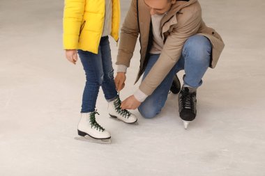 Genç baba buz patenlerinde bağcıklarını bağlıyor. Küçük kız buz pistinde.