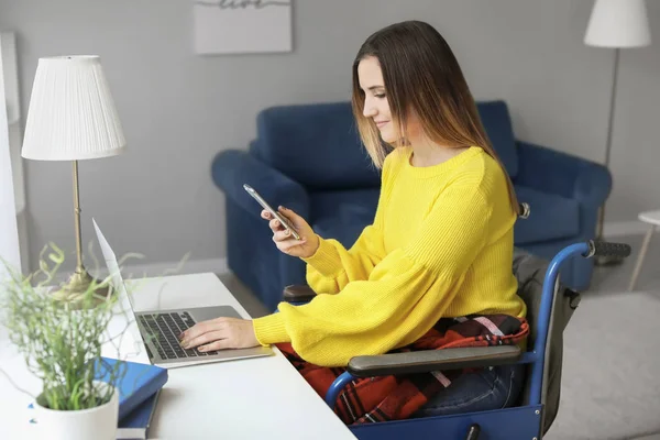 Mujer joven discapacitada en silla de ruedas usando portátil y teléfono móvil en casa — Foto de Stock