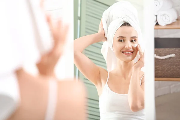 Krásná mladá žena nanášení krém na obličej před zrcadlem — Stock fotografie