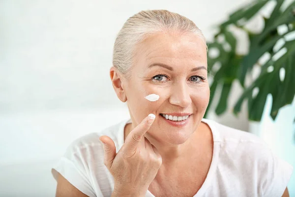 Hermosa mujer madura aplicando crema facial en casa — Foto de Stock