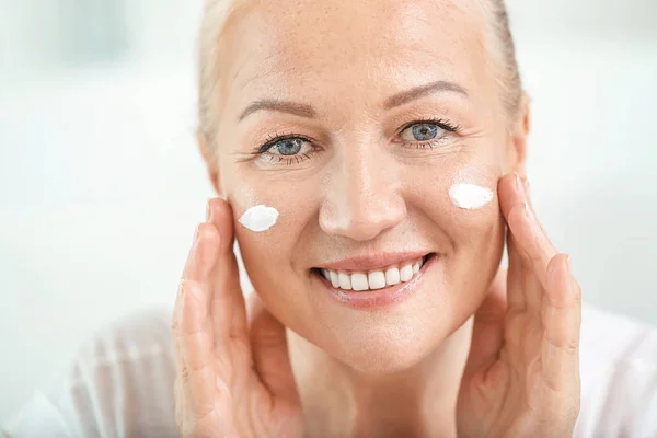 Mulher madura bonita aplicando creme facial em casa — Fotografia de Stock