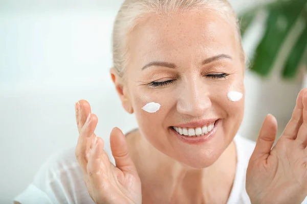 Mulher madura bonita aplicando creme facial em casa — Fotografia de Stock