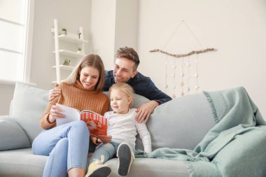 Mutlu genç aile evde kanepede otururken moda dergisi okuyor.