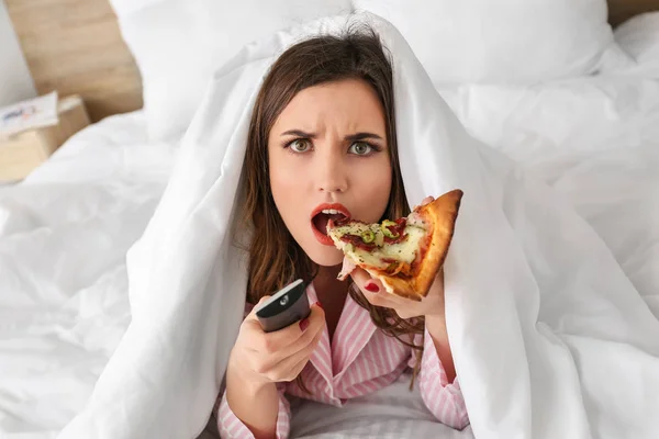 Ráno nespokojen mladá žena jíst chutnou pizzu při sledování televize v ložnici — Stock fotografie
