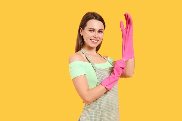 Young housewife putting on rubber gloves against color background — 스톡 사진