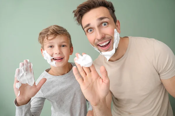 Funny father with son applying shaving foam onto their faces against color background — Stock Photo, Image
