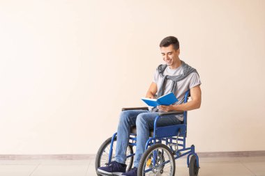 Handicapped young man in wheelchair reading book near color wall