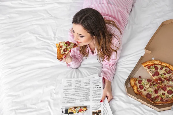Вранці красива молода жінка їсть смачну піцу і читає газету в спальні — стокове фото