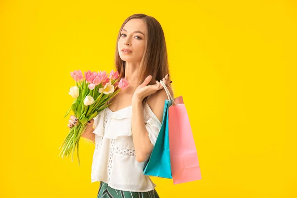 Piękna młoda kobieta z torbami na zakupy i kwiaty na tle koloru. Międzynarodowy Dzień Kobiet — Zdjęcie stockowe