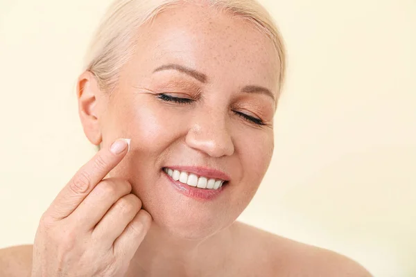 Beautiful mature woman applying facial cream against color background — 스톡 사진