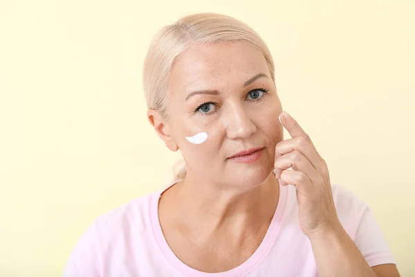 Mulher madura bonita aplicando creme facial contra fundo de cor — Fotografia de Stock