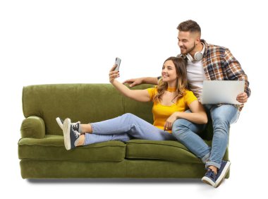 Dizüstü bilgisayarı ve cep telefonu olan genç bir çift beyaz arka planda koltukta otururken selfie çekiyor.