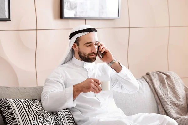 Hombre árabe guapo bebiendo café mientras habla por teléfono en casa — Foto de Stock