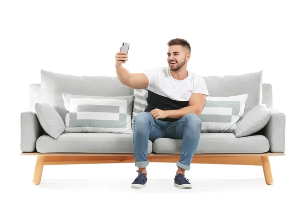 Jovem tomando selfie enquanto sentado no sofá contra fundo branco — Fotografia de Stock
