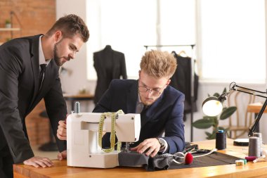 Genç erkek terzi ve meslektaşı atölyede dikiş makinesi üzerinde çalışıyorlar.