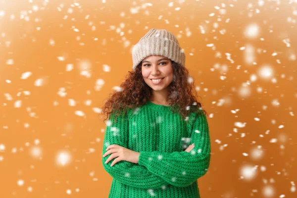 Young African-American woman in warm sweater and falling snow on color background — Stock Photo, Image