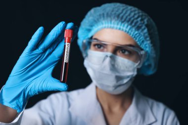 Koyu arkaplanda Coronavirus yazısıyla test tüpünde kan örneği tutan kadın doktor.