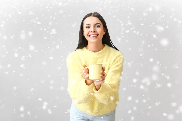 Mooie jonge vrouw in warme trui met thee en vallende sneeuw op lichte achtergrond — Stockfoto